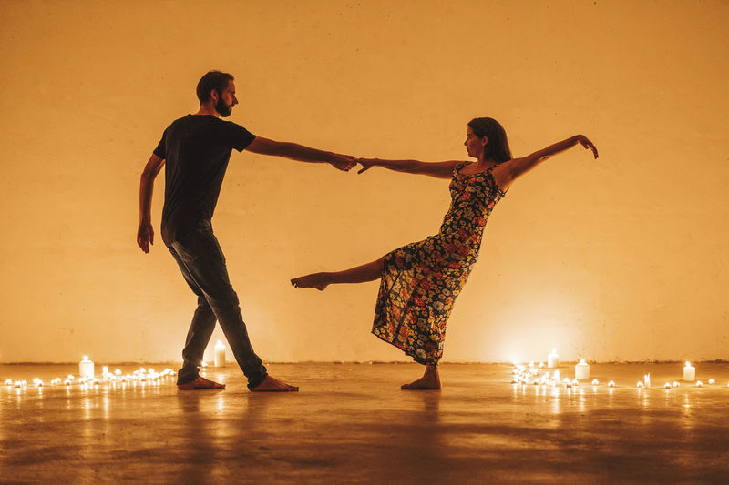 Couple holding hands while dancing by lit candles against wall in darkroom at home