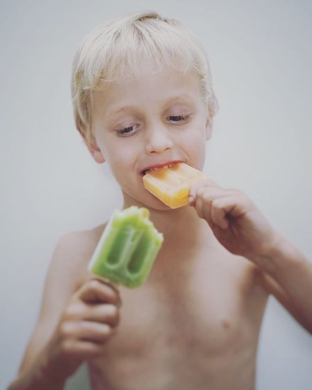 Close-up of shirtless boy eating popsicles while standing against white background