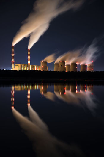Smoke emitting from factory with reflection on lake against sky at night