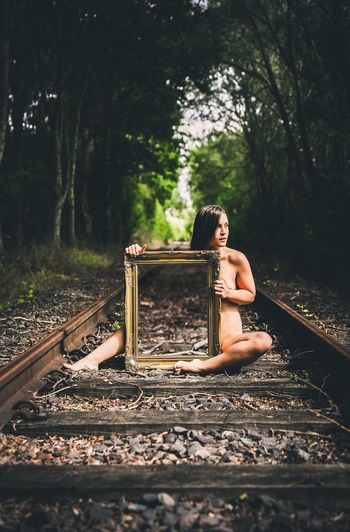 Full length of naked woman with mirror sitting on railroad track at forest