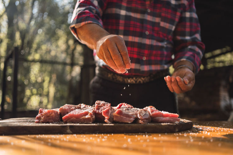 Crop unrecognizable man in checkered shirt putting salt on pieces of meat on wooden cutting board while preparing food in woods