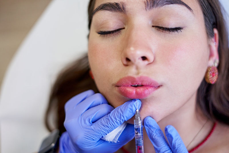 From above crop unrecognizable esthetician in latex gloves injecting collagen into lips of relaxed female client during appointment in beauty clinic