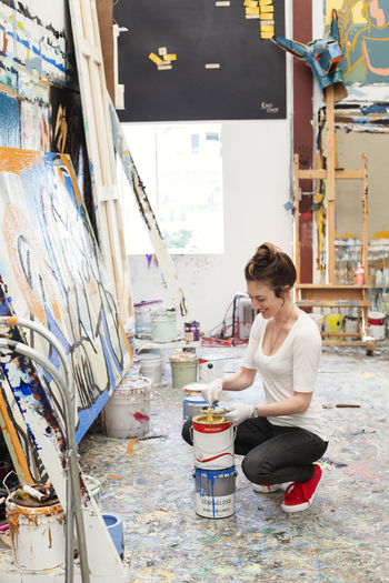 Happy artist crouching while dipping brush in paint can at art studio