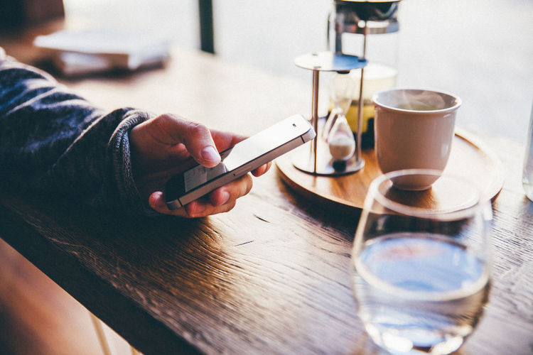 Cropped image of person using mobile phone by tea cup on table