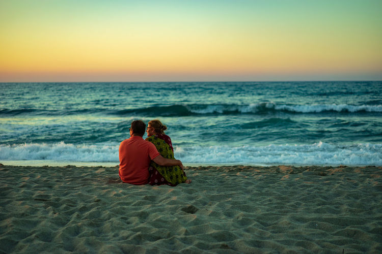 Rear view of couple sitting at beach against sky during sunset