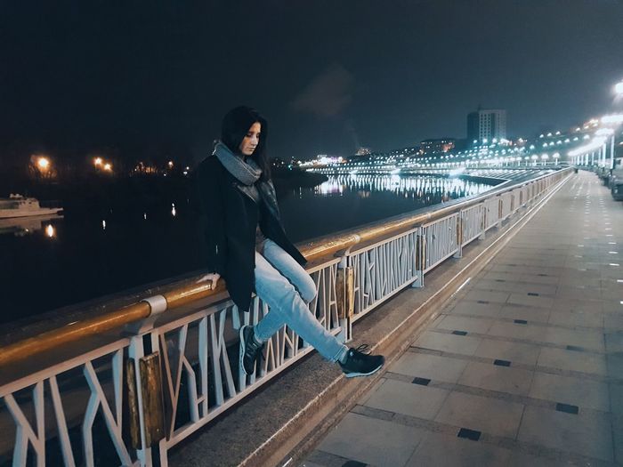 Young woman sitting on railing against river in city at night