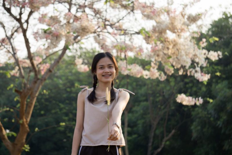 Portrait of young woman holding flower while standing against trees