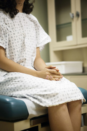 Midsection of woman with hands clasped sitting on bed in hospital