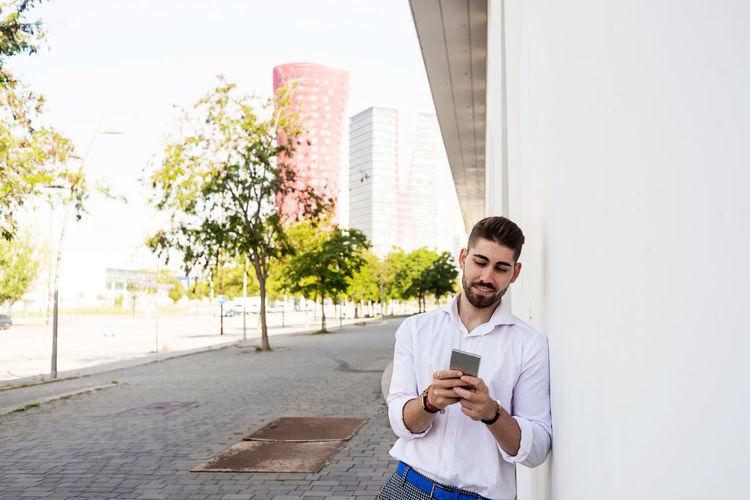 Young man using mobile phone standing against wall