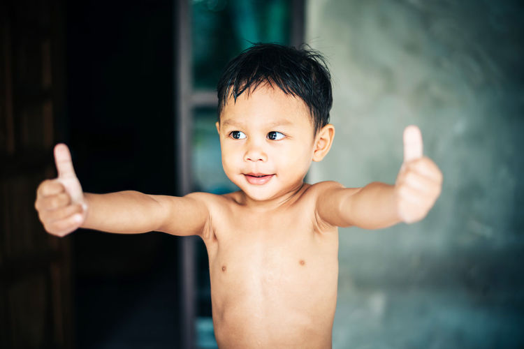 Shirtless boy showing thumbs up while standing at home