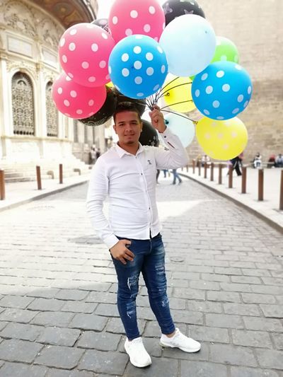 Full length portrait of young man holding colorful balloons while standing on footpath