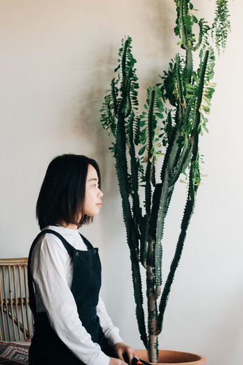 SIDE VIEW OF WOMAN LOOKING AT PLANT AGAINST WALL