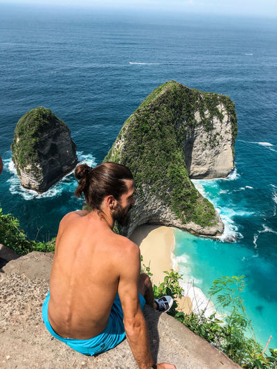High angle view of shirtless man sitting at cliff against sea