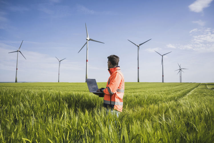 Engineer with laptop on wheat field looking at wind turbines