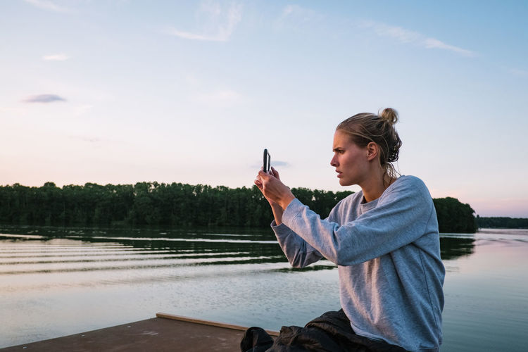 Side view of woman photographing while sitting on pier over lake against sky