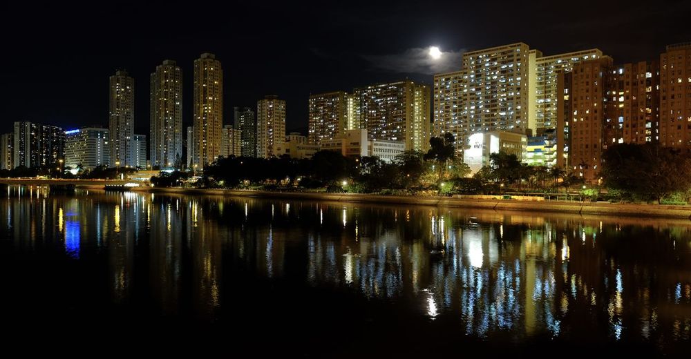 Illuminated buildings by lake against sky in city at night