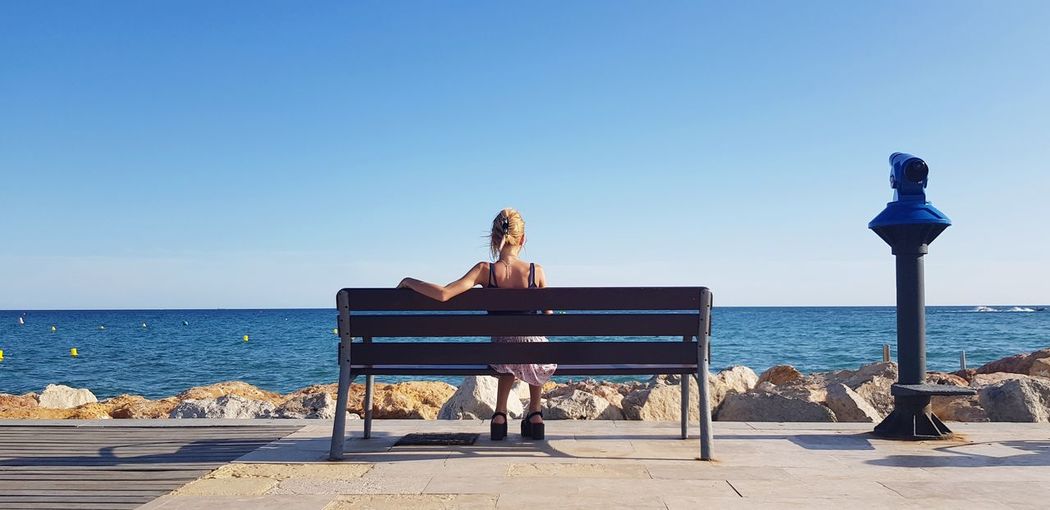 Rear view of woman sitting on bench by sea against sky