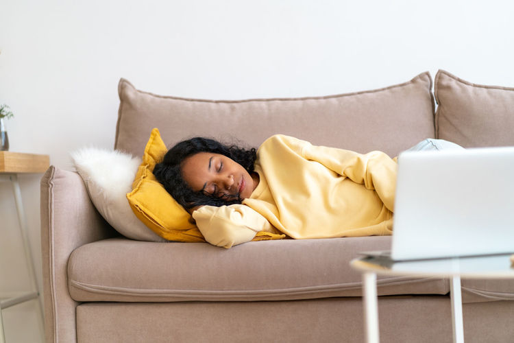 Bored african-american female sleeping on couch in living room while movie playing on laptop