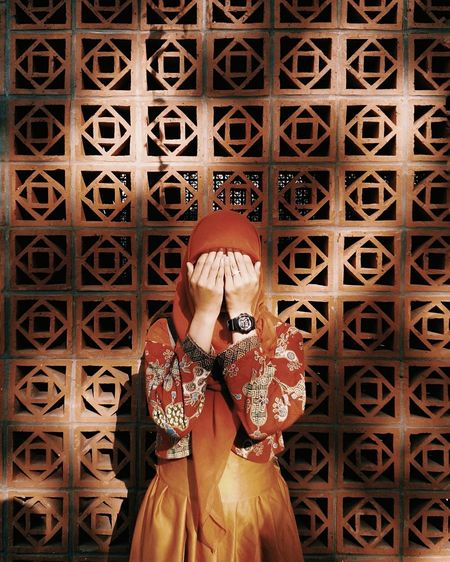 Woman covering her face with hands while standing against patterned wall