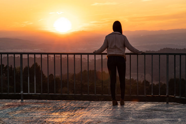 Rear view of woman standing by railing against sky during sunset