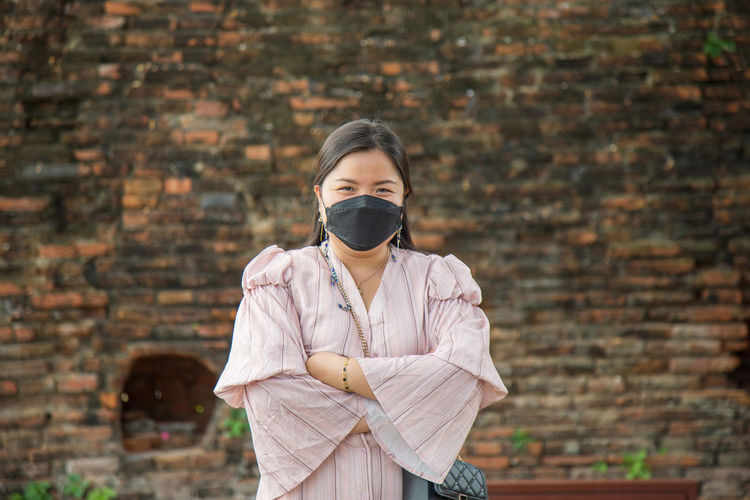 Asian woman in a pink dress crosses her arms wearing a face mask with an old brick wall background.