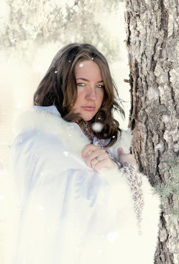 Portrait of young woman wearing fur coat while standing by tree during snowfall