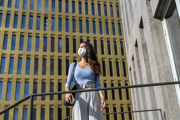 Young businesswoman in face mask looking away while standing against office building during coronavirus outbreak