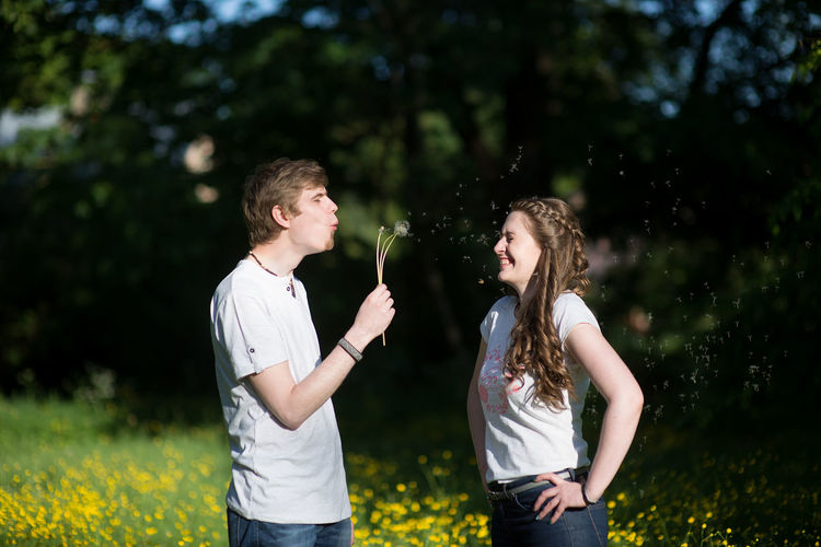 Man blowing dandelion seeds on cheerful girlfriend standing at park