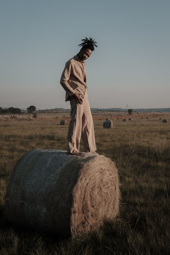 Full length portrait of young man standing on hay bale at agricultural landscape