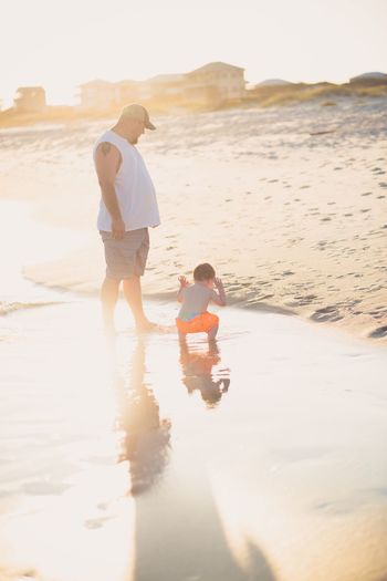 Father and son standing at beach against sky during sunset