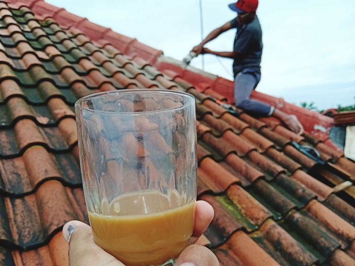 Cropped hand holding tea with man working on roof in background