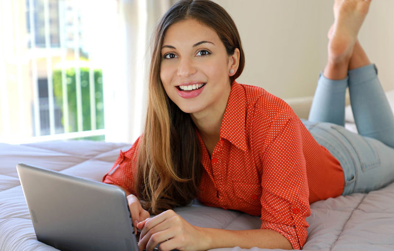 Portrait of smiling woman using laptop while lying on bed at home