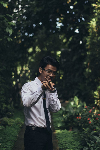 Portrait of smiling young man gesturing while standing on footpath against trees