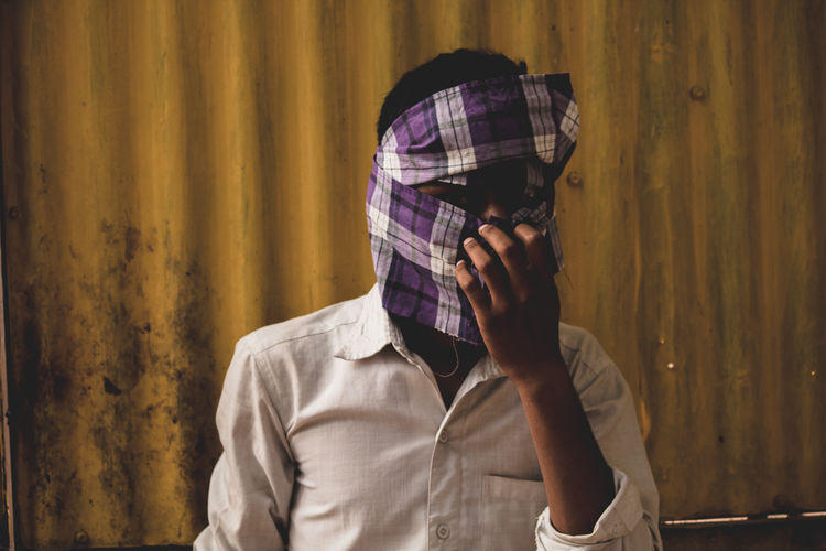 Portrait of teenage boy covering face with clothing while standing against wall