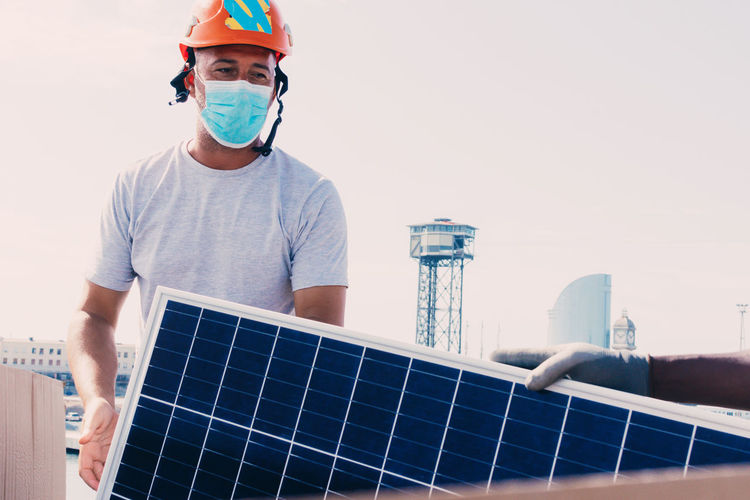 Workman in helmet and protective mask installing solar battery in industrial area of contemporary plant