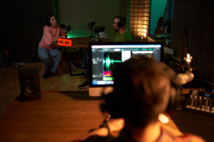 Back view of unrecognizable male sound director sitting at table with computer during podcast recording with host and female singer in dark studio