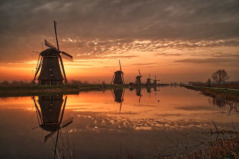 Traditional windmill by lake against cloudy sky during sunset