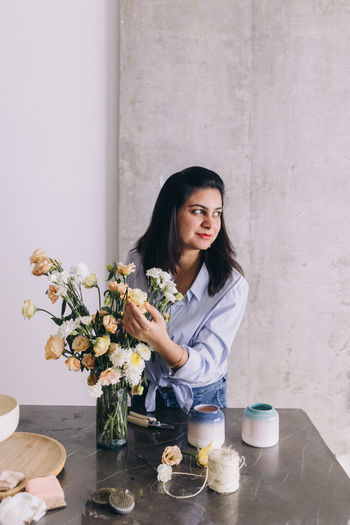 Portrait of smiling young woman against wall looking away and holding a flower bouquet 