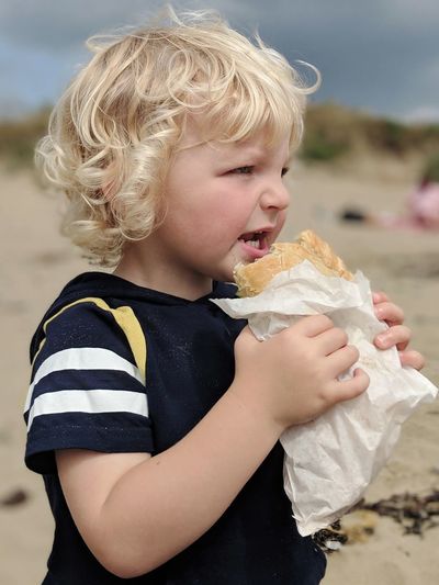 Cute boy eating food while standing at beach against cloudy sky