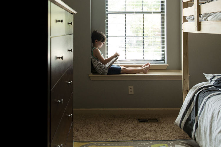Boy reading book while sitting on window sill