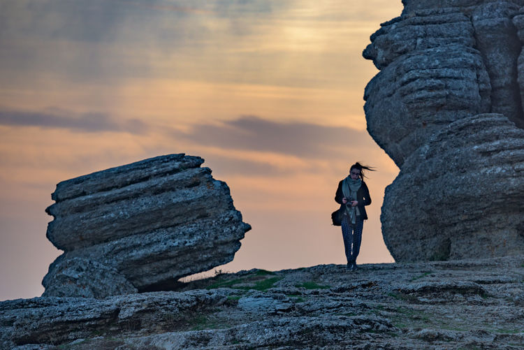 Woman with camera standing by rock formations at el torcal de antequera against cloudy sky during sunset