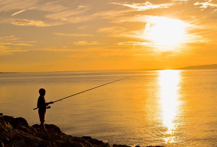 Silhouette boy fishing in sea against sunset sky