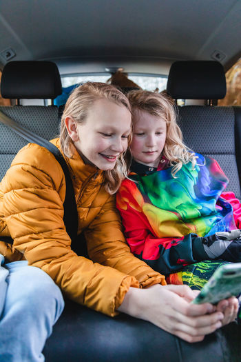 Excited sisters sitting in car and playing interesting game on mobile phone together