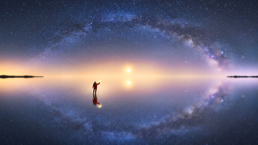 Silhouette of anonymous man standing on reflection surface of water and reaching out to starry colorful night sky with milky way