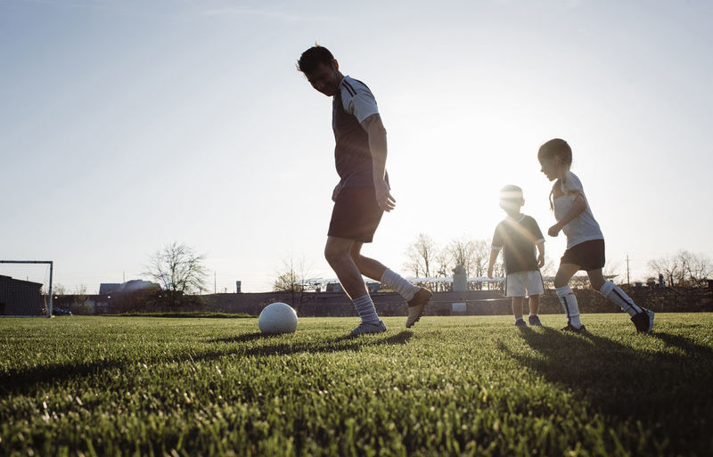Low angle view of man playing soccer with children on grassy field against clear sky during sunset