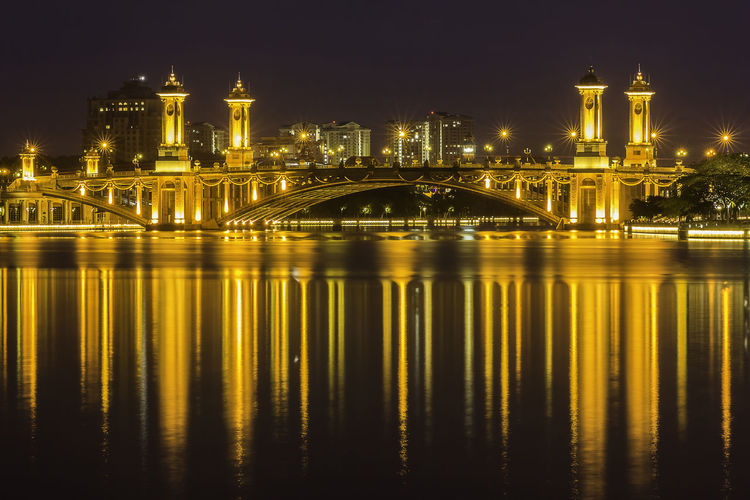 Illuminated arch bridge reflecting on river against sky at night