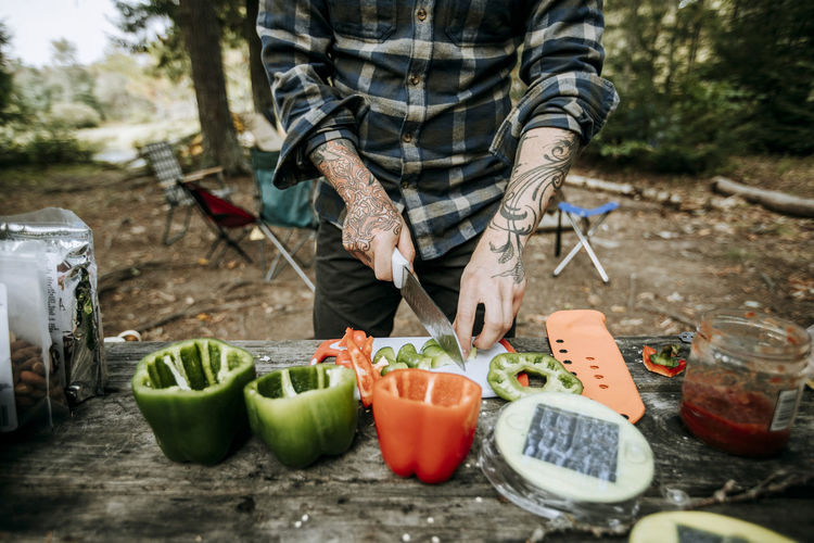 Man with tattoos chops vegetables on wood table while camping in woods