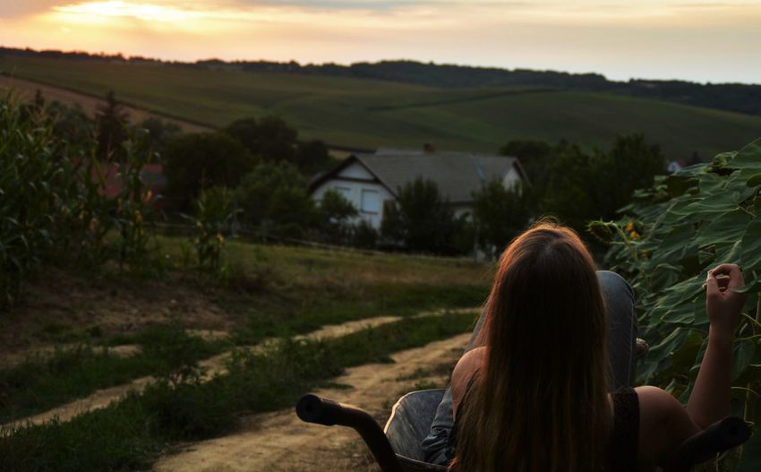 Rear view of woman sitting in wheelbarrow on dirt road against mountain during sunset