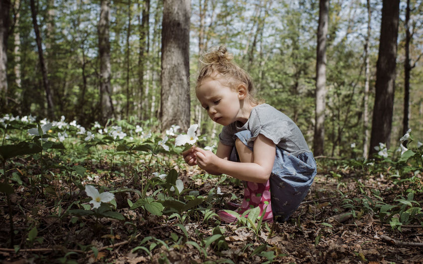 Side view of cute girl looking at white flower while crouching in forest