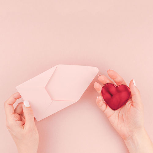 Cropped hands of woman holding envelope and heart shape over pink background